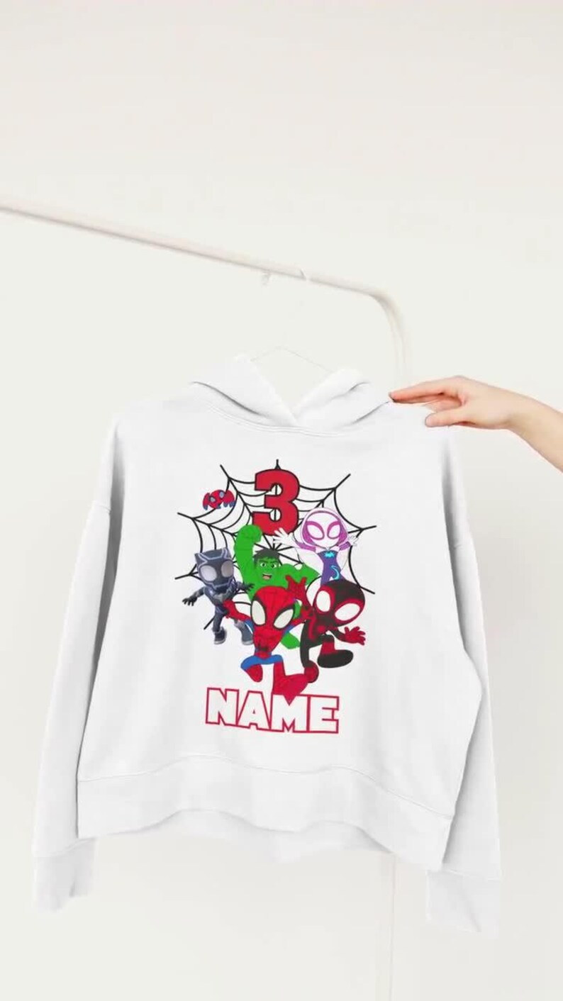 May include: White hooded sweatshirt featuring a colorful graphic of several cartoon superheroes, a red number 3, and the word "NAME" in red block letters. The design is set against a spiderweb background.