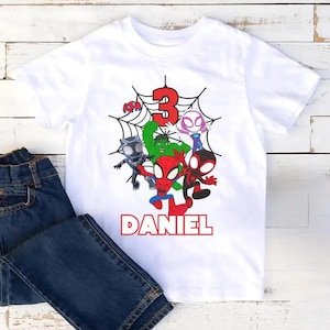 May include: White t-shirt featuring a colorful graphic of several cartoon superheroes, a red number 3, and the name "DANIEL" in red block letters. The shirt is paired with a pair of blue denim jeans.