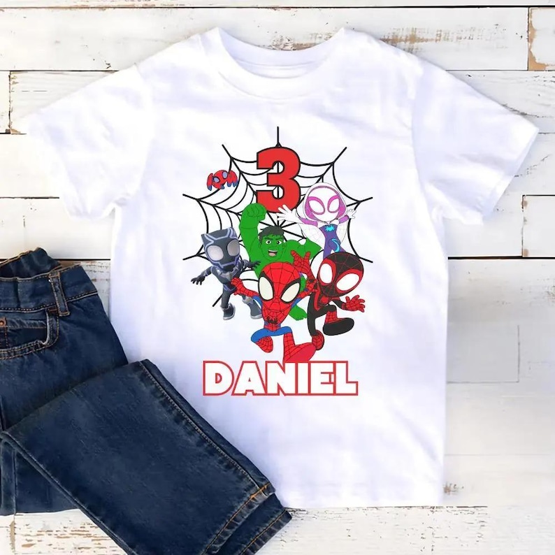 May include: White t-shirt featuring a colorful graphic of several cartoon superheroes, a red number 3, and the name "DANIEL" in red block letters. The shirt is paired with a pair of blue denim jeans.