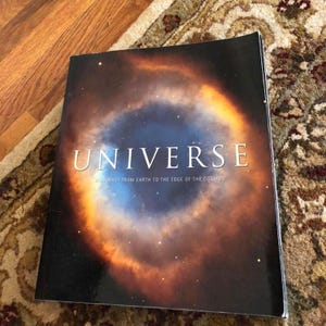 Puede incluir: Un libro titulado "UNIVERSE" con una imagen vibrante de nebulosa en la portada. La imagen presenta una formación de nubes cósmicas azules y naranjas. El subtítulo dice "Journey from Earth to the Edge of the Cosmos."
