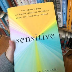 May include: A book titled "sensitive" is held in a person's hand. The cover features a gradient of colors, including yellow, blue, and green. The title is in large, black letters. The book is about highly sensitive people.