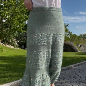 Può includere: Una gonna midi in maglia color verde salvia con vita a coste, un motivo a maglia aperta testurizzato e paillettes iridescenti sparse su tutto il tessuto. La gonna presenta un orlo svasato e una consistenza morbida e soffice, abbinata a una camicetta bianca.
