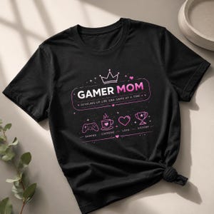 May include: Black t-shirt with a pink graphic that reads "GAMER MOM" with a crown and gaming-related icons. The text "LEVELING UP LIFE ONE GAME AT A TIME" is also included. The shirt features a crew neck and short sleeves.