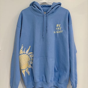 May include: A light blue hoodie with a front pocket and drawstring hood. The left sleeve features a yellow and white sun graphic. The text "BE THE LIGHT" is printed on the upper right chest.