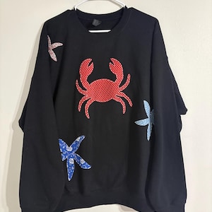 Puede incluir: Sudadera negra con un aplique de cangrejo rojo con lunares blancos. La sudadera también presenta tres apliques de estrellas de mar en tonos azules y rosas. La sudadera está colgada de una percha blanca.