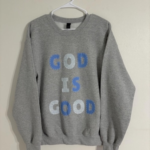 May include: A gray sweatshirt with the words "GOD IS GOOD" in blue and white letters. The sweatshirt is hanging on a white hanger against a white wall. The letters are stitched onto the front of the sweatshirt.