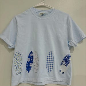 May include: Pale blue t-shirt featuring five oval patches in assorted blue and white floral and gingham designs. The garment is suspended on a white hanger against a light grey wall.