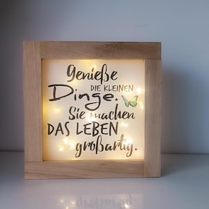 Könnte beinhalten: Holzgerahmte Leuchtbox mit den Worten "Genieße die kleinen Dinge. Sie machen DAS LEBEN großartig." in schwarzer Schrift. Auf der rechten Seite befindet sich ein kleiner grüner Schmetterling. Die Box ist mit kleinen, warmweißen Lichtern beleuchtet.