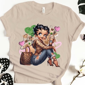 May include: Beige t-shirt featuring a cartoon illustration of a woman with dark hair, wearing a brown hoodie and ripped jeans. She holds a coffee drink and sits next to a brown handbag. Pink and green heart shapes are in the background.