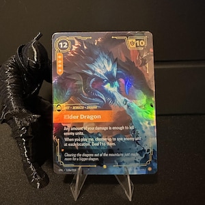 Könnte beinhalten: Holografische Sammelkarte mit einem Elder Dragon. Die Karte zeigt eine blaue und violette Drachenillustration mit dem Text "Elder Dragon". Die Karte wird auf einem transparenten Kunststoffständer präsentiert.