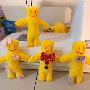 May include: Four yellow plush dolls with embroidered red eyes and mouths. One doll is plain, one has pink ribbon bows and flowers, one features a red and white polka dot bow tie with two buttons, and one wears a purple bow tie with a small pearl accent.