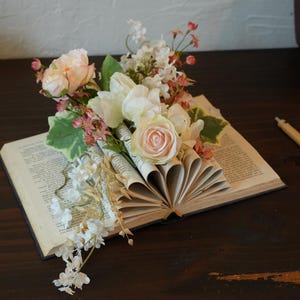 May include: An open book with pages arranged to display flowers. The arrangement includes light pink and white roses, greenery, and delicate white flowers. A pen rests nearby on a dark wooden surface.