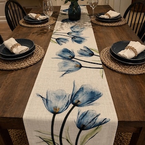Op de afbeelding: Een houten eettafel gedekt voor een maaltijd, met een bloemen tafelloper met blauwe tulpenontwerpen. De tafel is gedekt met donkerblauwe borden, witte servetten en geweven placemats. Wijnglazen en een vaas met groen maken de elegante setting compleet.