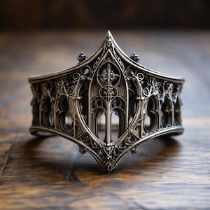 Op de afbeelding: Een sierlijke zilverkleurige manchetarmband met een gotisch architectonisch ontwerp. De armband heeft ingewikkelde details van bogen, torens en decoratieve elementen. De manchet wordt op een houten oppervlak gepresenteerd.