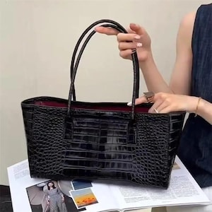 May include: A black tote bag with a glossy crocodile skin texture and two thin handles. The interior of the bag is lined with a deep burgundy fabric. The bag has a rectangular shape with a structured base and a wide opening at the top.