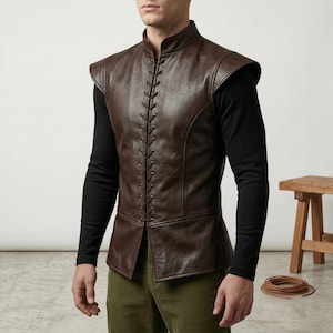 May include: A brown leather vest with a high collar and shoulder pads. The vest features a lace-up front and is worn over a black long-sleeved shirt. The person is also wearing green corduroy pants. A wooden stool and a coil of rope are in the background.