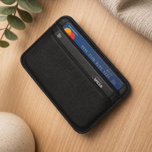 Pode incluir: Um porta-cartões de couro preto com duas ranhuras, contendo um cartão Mastercard e um cartão Visa. O Mastercard é azul, laranja e vermelho, enquanto o cartão Visa é preto e branco. O porta-cartões é retangular e tem um design minimalista.