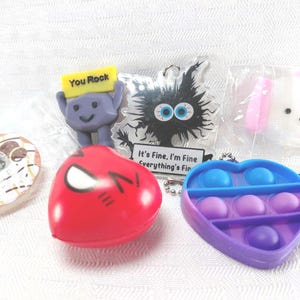 May include: A collection of colorful novelty items, including a red heart-shaped toy, a purple heart-shaped pop-it toy, a donut, a cartoon character holding a sign that says "You Rock", a black and white cartoon character, and a white dog with pink ears.