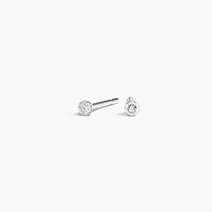 May include: A pair of silver-toned stud earrings, each featuring a single round, clear, faceted stone set in a metallic base. One earring is shown from the side, revealing the straight post, while the other is positioned to display the front face.