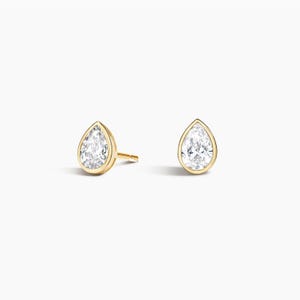 May include: A pair of stud earrings featuring pear-shaped clear stones set in yellow gold bezels. Each earring has a smooth, polished gold rim surrounding the faceted stone, with a post backing visible on the left earring against a plain background.