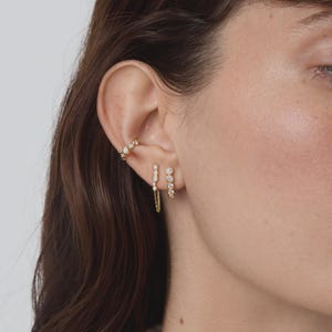 May include: A gold ear cuff featuring a row of small round bezel-set clear stones, paired with two gold stud earrings. One stud features a vertical row of stones with a delicate dangling chain, while the other is a simple vertical row of stones.