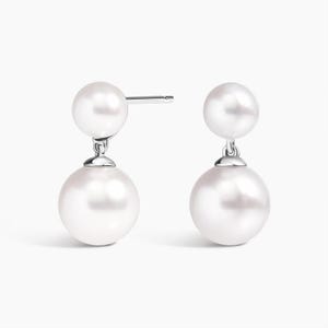 Pode incluir: Um par de brincos pendentes com duas pérolas esféricas brancas de tamanhos diferentes. A pérola menor fica no topo, perto do pino prateado, enquanto a pérola maior pende abaixo, conectada por um pequeno elo de metal prateado.