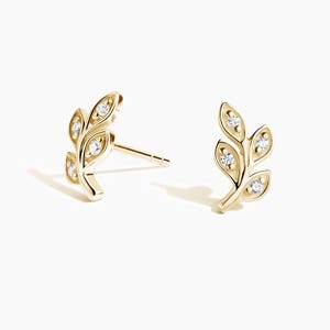 May include: A pair of gold stud earrings shaped like delicate leaves, each featuring three small, clear, round-cut stones embedded within the metal branches. The earrings have a polished finish and include a standard post backing for secure attachment.