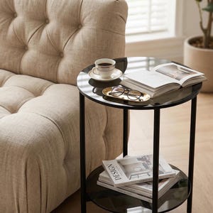 May include: A two-tiered black side table with a glass top and a lower shelf. A cup of coffee, a book, and a tray with glasses rest on the top. Magazines with the word "DESIGN" are on the lower shelf. The table is next to a beige sofa.