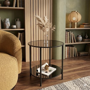 May include: A modern black side table with a round, smoked glass top and a lower shelf. The table has a minimalist design with a metal frame. A clear glass vase with dried flowers sits on top. The table is 20 inches in diameter.