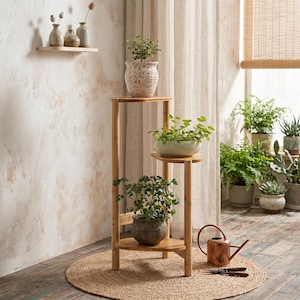Op de afbeelding: Een bamboe plantenstandaard met drie niveaus met potplanten. De standaard biedt plaats aan een verscheidenheid aan planten in decoratieve potten. Een koperen gieter en schaar liggen op een ronde jute mat.