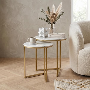 May include: Two round nesting tables with white marble tops and gold metal frames. A mug, decorative items, and a vase with dried flowers sit on the tables. The tables are a stylish addition to any living room.