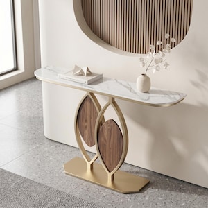 May include: A contemporary console table with a white marble top and a gold-coloured metal frame. The base has two wooden panels within an open, leaf-shaped design. A small vase and a book are on the table.