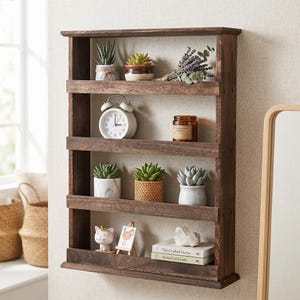 Puede incluir: Estantería de pared de madera marrón oscuro con cuatro estantes horizontales. Los estantes exhiben varios artículos, incluyendo suculentas en macetas, un despertador blanco, una vela, un ramo de flores secas, una figura de gato y libros.