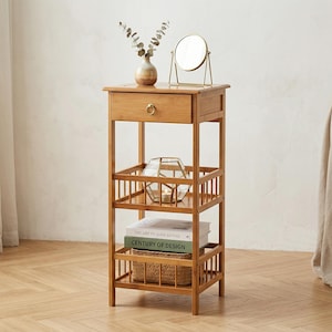 Puede incluir: Una mesa auxiliar de bambú marrón claro con un cajón, estantes y una cesta. El estante superior contiene un espejo pequeño y un jarrón con vegetación. Los estantes inferiores contienen un portavelas geométrico y libros. La mesa mide 71 cm de alto.