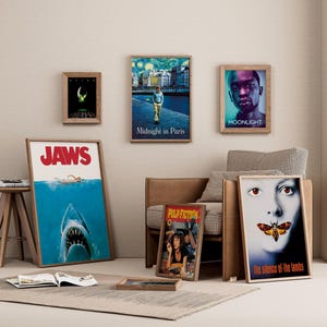 May include: A collection of framed movie posters, including "Jaws," "Midnight in Paris," "Moonlight," "Pulp Fiction," and "The Silence of the Lambs." The posters are displayed on a neutral-toned wall, with a rug and small wooden table.