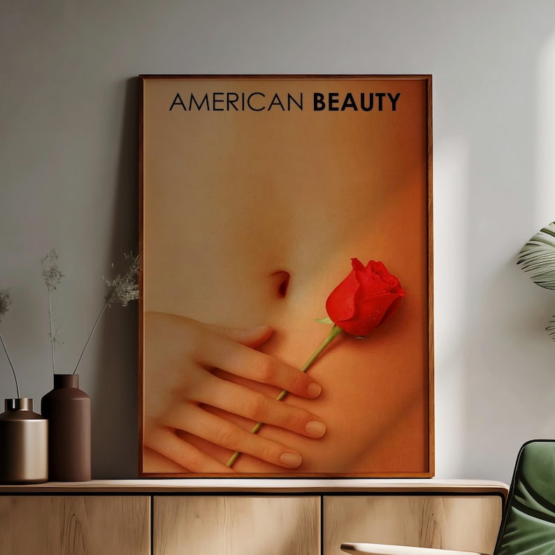 May include: A framed print featuring the text "AMERICAN BEAUTY" above an image of a red rose held near a person's abdomen. The image has a warm, orange and brown color palette.