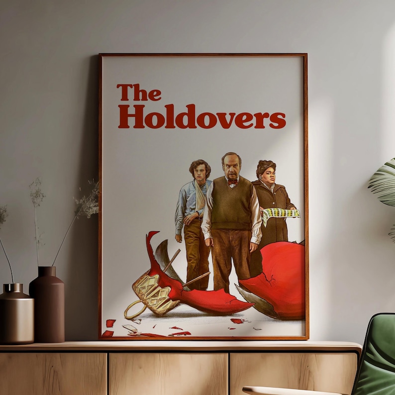 May include: A framed movie poster for "The Holdovers" featuring three people and a shattered ornament. The title is in red, with the figures standing above a broken red ornament. The poster is in a brown frame.