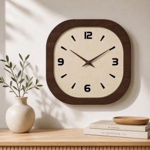 May include: A square wall clock with rounded corners and a dark brown wooden frame. The clock face is a light beige colour with black numerals and hands. The clock is mounted on a light-coloured wall.