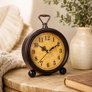 May include: A vintage-style, round analog clock with a dark brown metal frame and a handle. The clock face is a light tan color with black numerals and hands. It has two black ball feet and sits on a wooden surface.