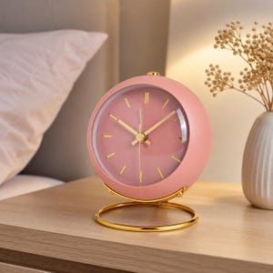 Puede incluir: Un despertador analógico redondo de color rosa empolvado con un acabado mate liso. La esfera tiene un fondo rosa a juego, marcadores de hora dorados finos y manecillas doradas delgadas. Se apoya sobre un soporte circular de metal dorado.
