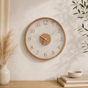 Peut inclure: Une horloge murale ronde avec un cadre en bois clair et un cadran en verre transparent. L'horloge présente un fond clair avec des chiffres noirs et des aiguilles en bois clair. Le centre de l'horloge est un cercle en bois plus foncé. Un objet décoratif pour la maison.