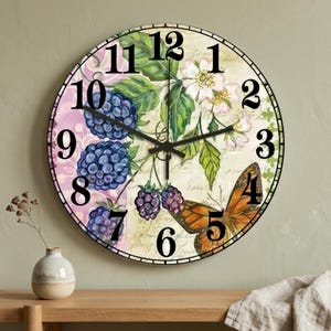 Pode incluir: Um relógio de parede redondo com um design floral e borboleta. O mostrador do relógio apresenta um fundo creme com números pretos, uma borboleta e ilustrações de amoras e flores. Os ponteiros do relógio são pretos.