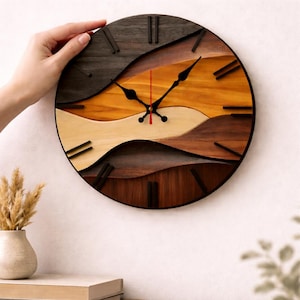 May include: A round wooden wall clock with a layered design. The clock face features a wave-like pattern in shades of brown, tan, and orange. Black hands and hour markers contrast against the lighter wood tones. The clock is approximately 12 inches in diameter.