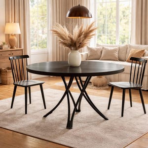 Puede incluir: Una mesa de comedor redonda de color marrón oscuro con una base de metal negro, rodeada por dos sillas de madera negras. Un jarrón blanco con hierba de la pampa seca está sobre la mesa. La habitación tiene una paleta de colores neutros con una alfombra y un sofá.