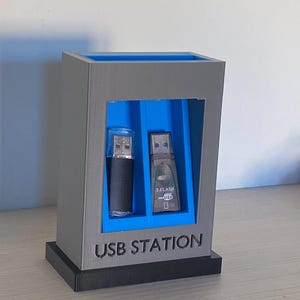 Può includere: Una stazione USB rettangolare grigia e blu con base nera. L'unità presenta due slot verticali che contengono una chiavetta USB nera e una chiavetta USB T-FLASH. Il testo USB STATION è impresso sulla parte anteriore della cornice grigia.