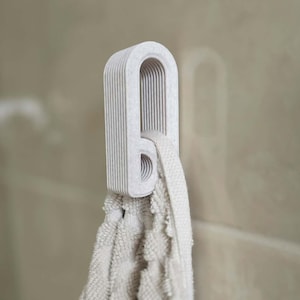 May include: A light beige towel hangs from a matching wall hook. The hook has a unique, layered design with a curved top and a circular opening. The towel is made of a soft, textured material, creating a spa-like aesthetic.