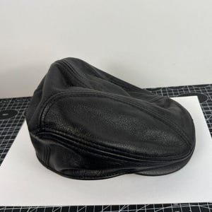 May include: A black leather flat cap featuring a textured surface with visible stitched seams across the crown and a short, rounded brim. The cap has a structured yet flexible form, showcasing the natural grain of the material throughout.