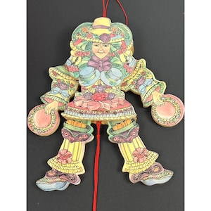 May include: A flat, articulated wooden hanging ornament shaped like a person wearing a wide-brimmed hat, ruffled sleeves, and layered clothing in pastel shades of yellow, pink, and blue, holding a small circular object in each hand with a red string.