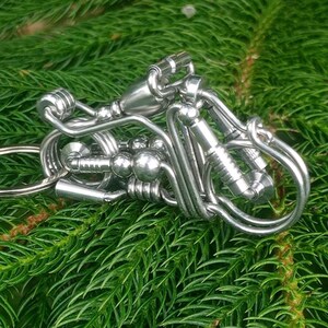 Könnte beinhalten: Ein silberfarbener Metall-Schlüsselanhänger in Form eines Motorrads, gefertigt aus gebogenem Draht, kleinen zylindrischen Komponenten und kugelförmigen Perlen. Das Design verfügt über einen runden Ring auf der linken Seite und Drahtdetails.