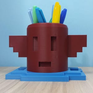 May include: A burgundy 3D-printed pen holder, resembling a pixelated face with rectangular cutouts for eyes and mouth. It features wing-like extensions and a blue base. The holder contains several colourful pens.